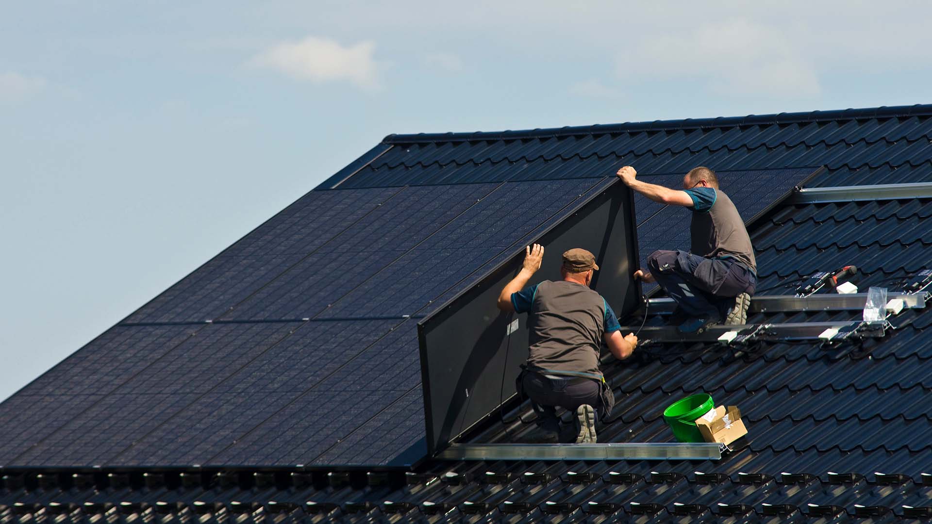 Two workers install solar panels on top of a house.